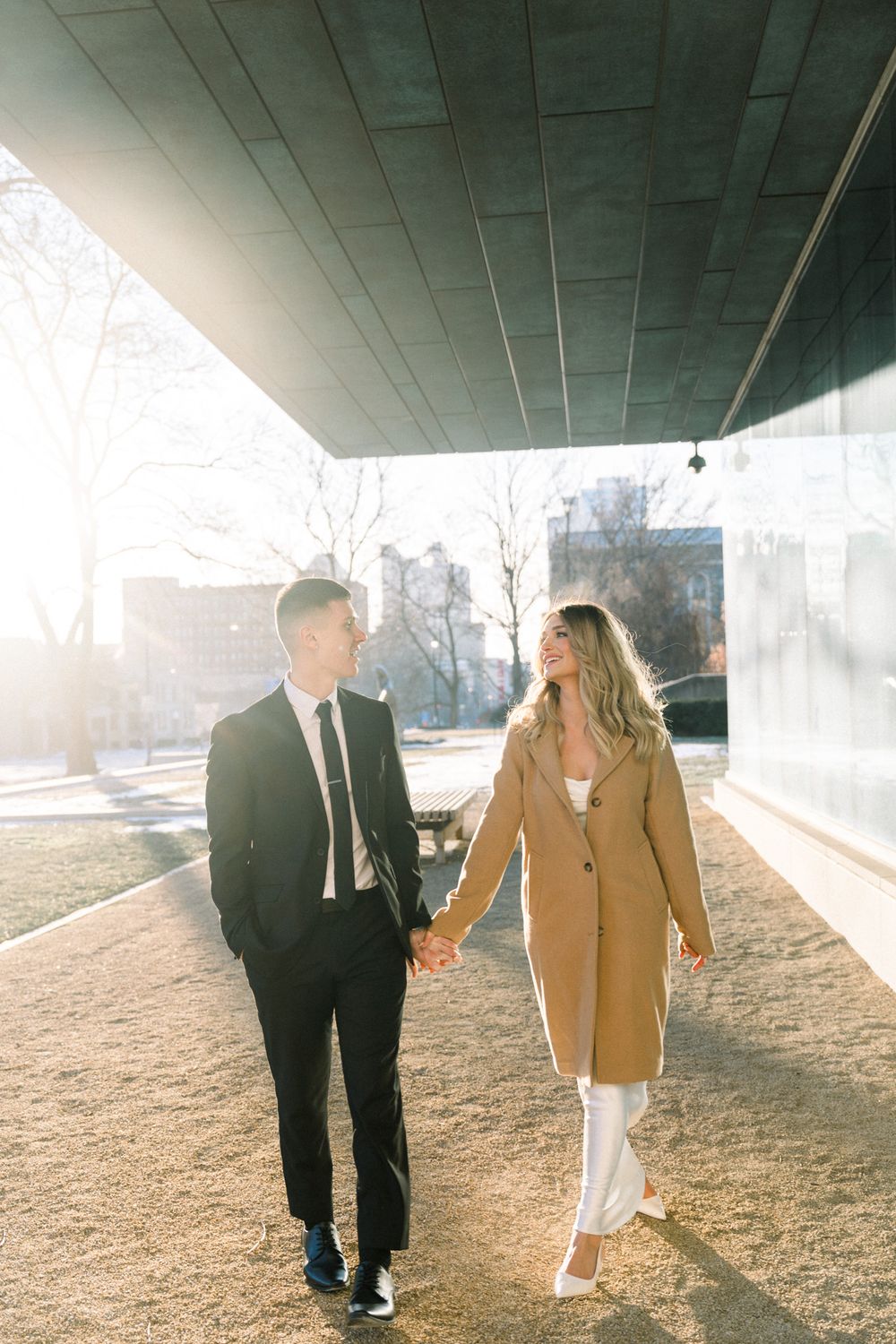 Sunlit couple holding hands while walking under concrete overhang in formal winter outfits.