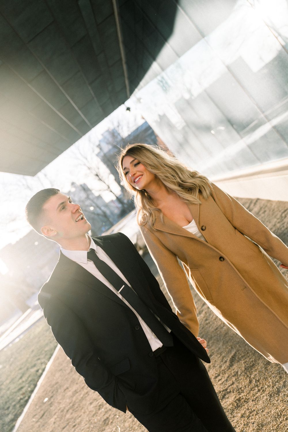 A couple shares a candid moment outdoors, with sunlight streaming through as they smile and laugh together.