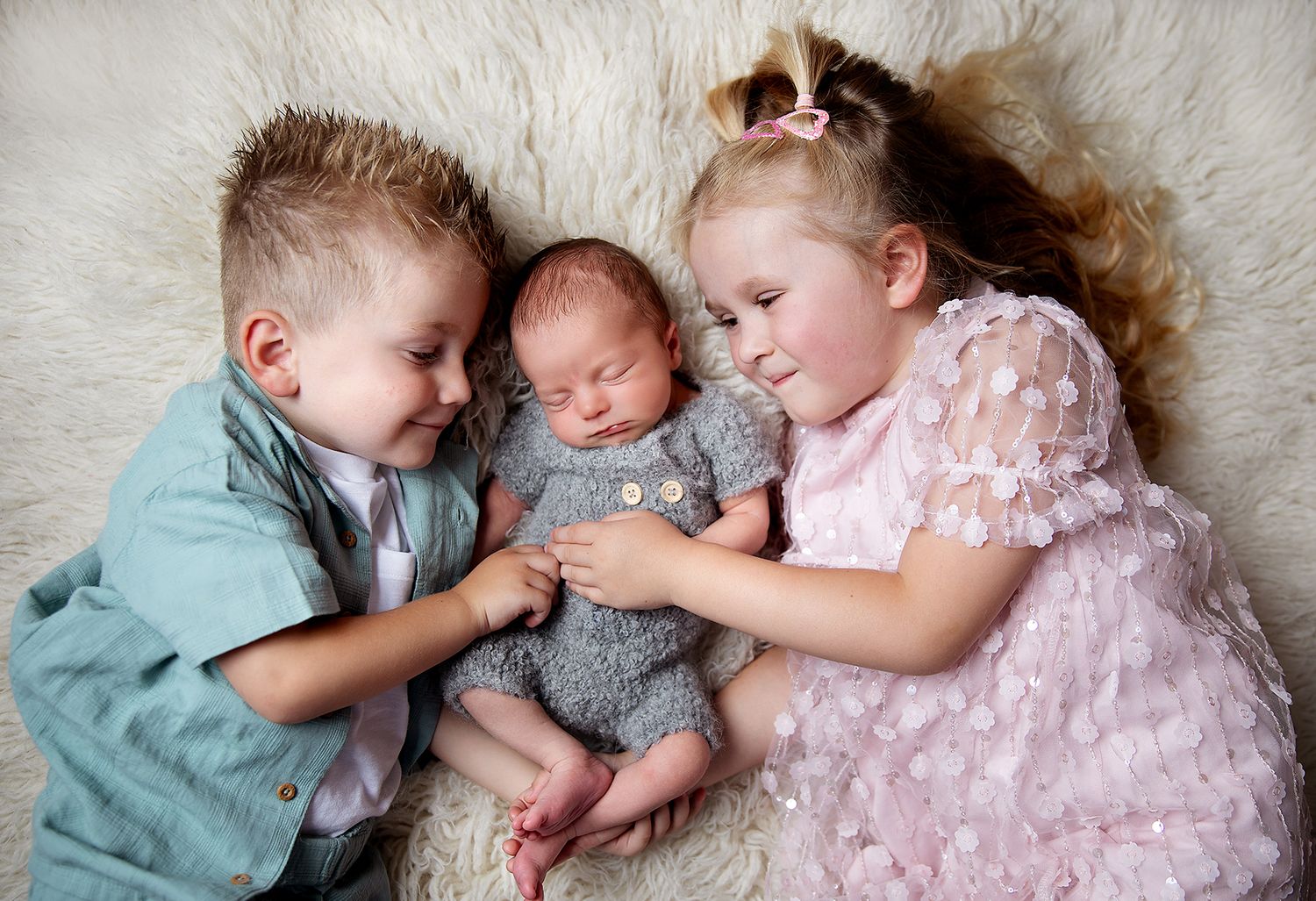 Three siblings lying together on a white fluffy rug, wearing blue and pink outfits as they share a sweet moment. During a newborn session