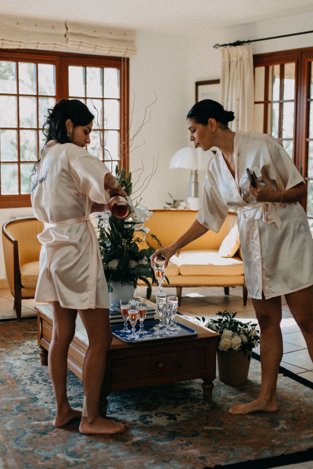 Two people in white robes getting ready on wedding day morning in a living room with floral arrangements.