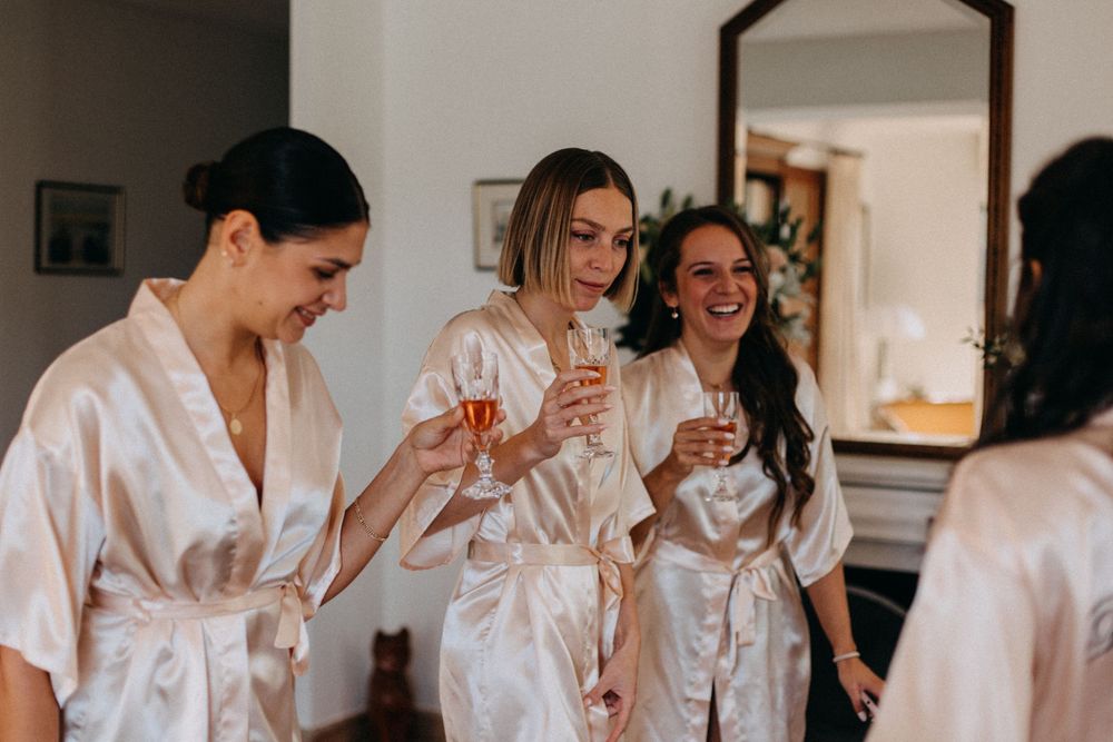 Wedding party bridesmaids in matching white satin robes share a joyful moment with drinks during preparations.