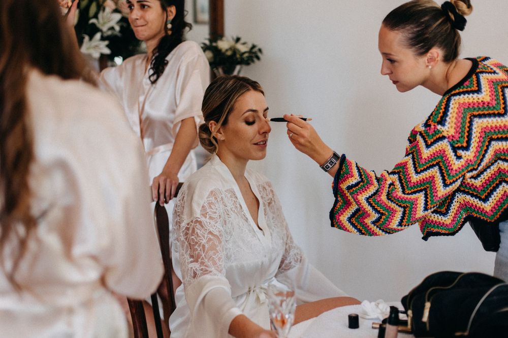 Makeup artists prepare a bride in a white lace robe during wedding day preparations.