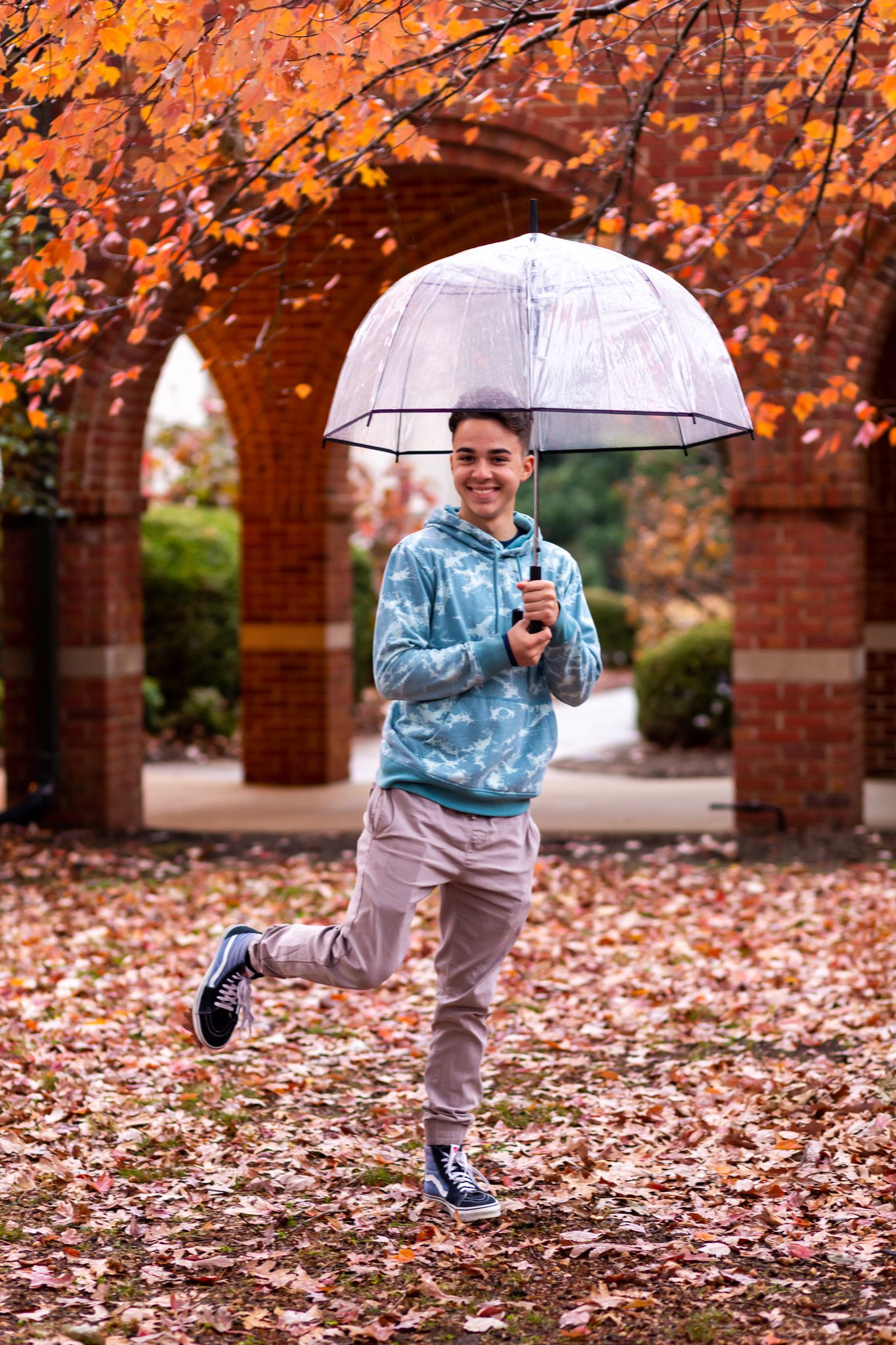 male teen holding clear umbrella with one foot kicked up behind. Silly posing for senior pictures with MegMc Photography, Medina, TN