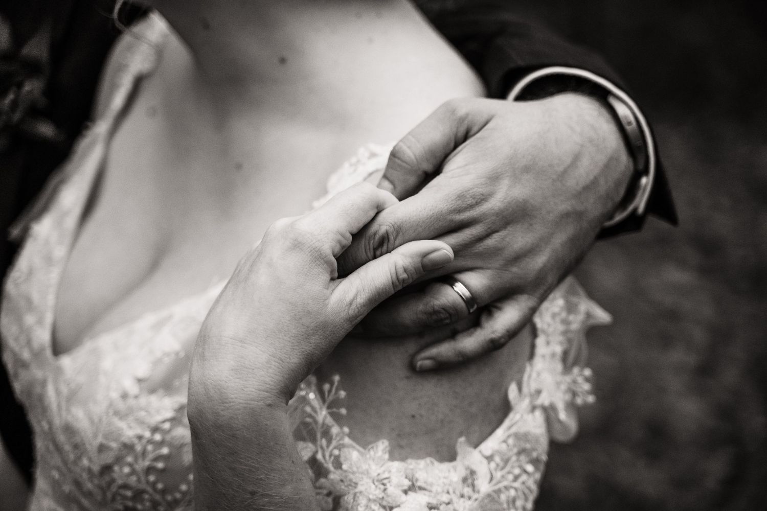 A black and white closeup of hands wearing wedding rings and lace details from a bridal gown.