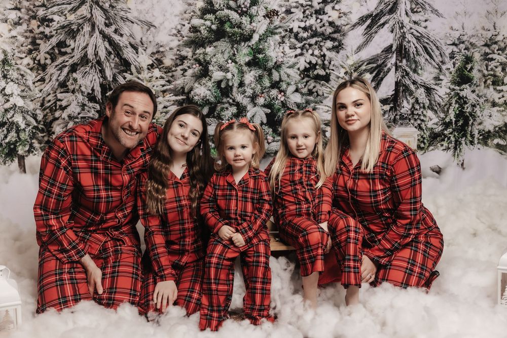 Family wearing matching red plaid pajamas poses together in front of snowy evergreen trees for winter holiday photos.