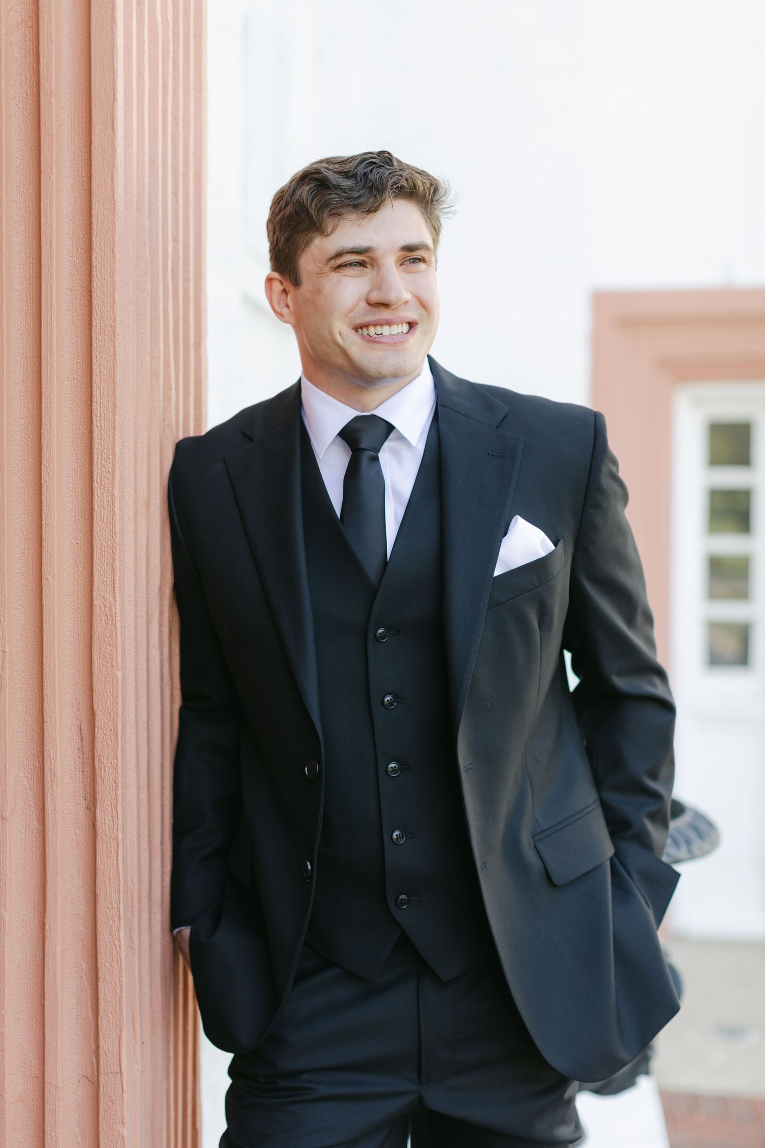 A young person in a classic black three-piece suit and tie leans against a coral-colored pillar while smiling.