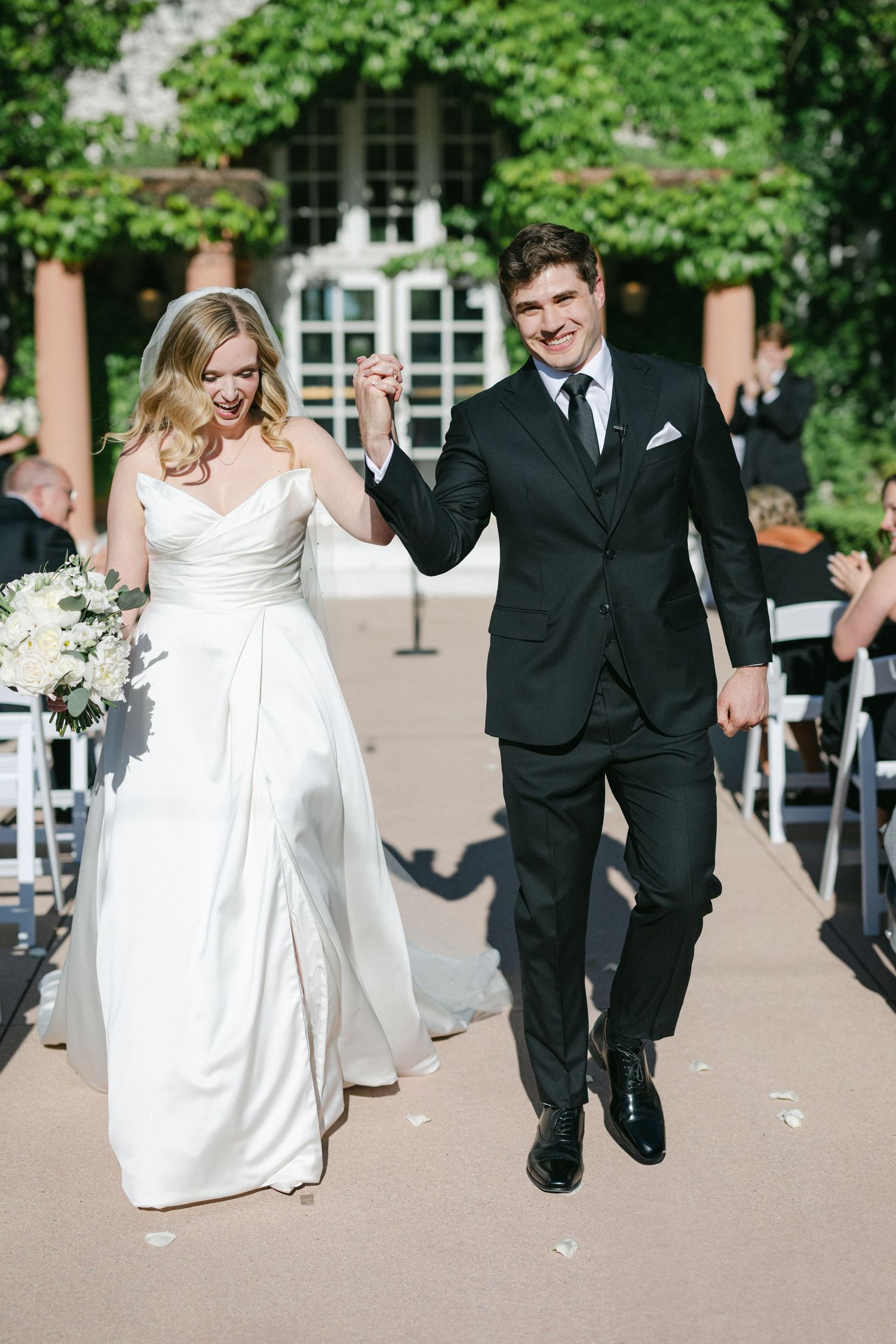 Four sequential photos show a couple in wedding attire playfully running and dancing together on a tree-lined path.