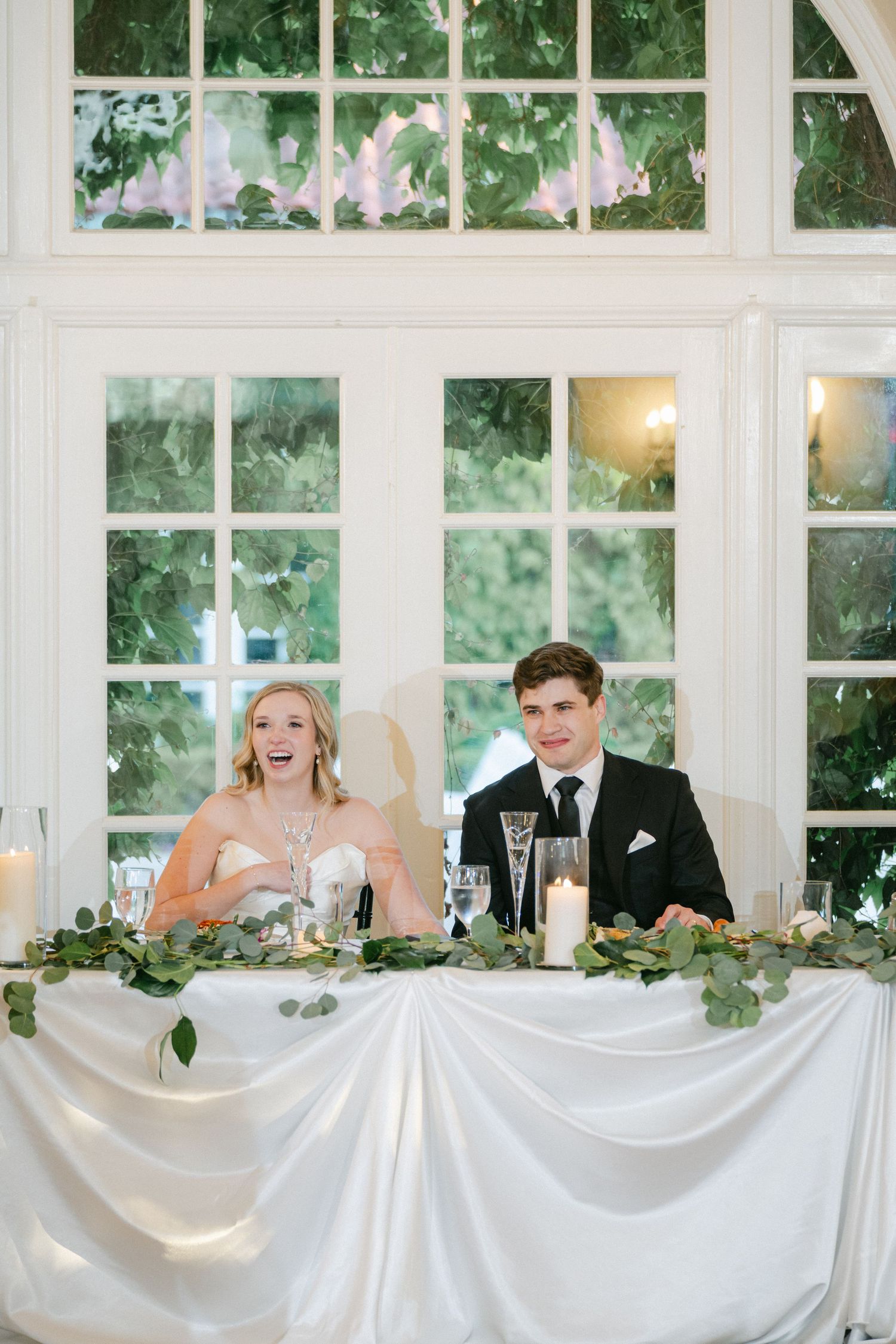 White draped wedding reception head table adorned with greenery garland and glass decor in elegant sunlit venue.