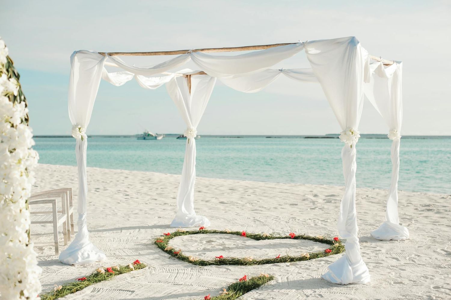 White fabric canopy setup on a tropical beach with a heart made of flowers in the sand and turquoise ocean waters. Wedding Photo & Video.