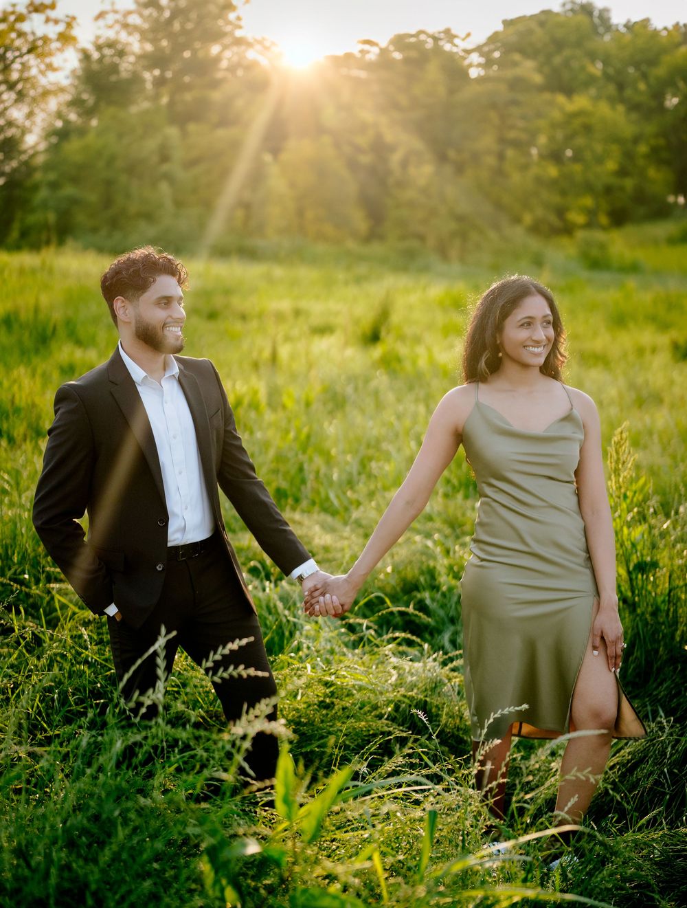 A couple holds hands in a sunlit meadow during golden hour as sun rays stream through trees in the background.