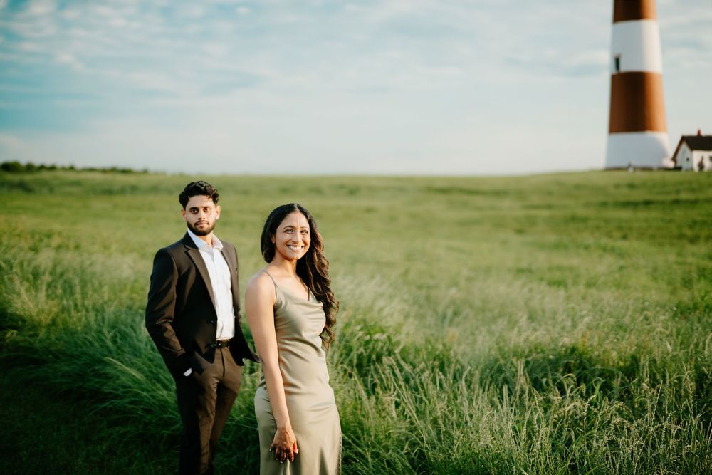 A couple stands in a green field near a striped lighthouse on a sunny summer day.
