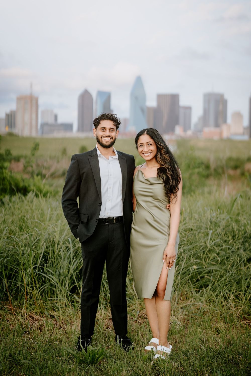 A couple poses together in formal attire on a grassy hill with a city skyline visible in the background.