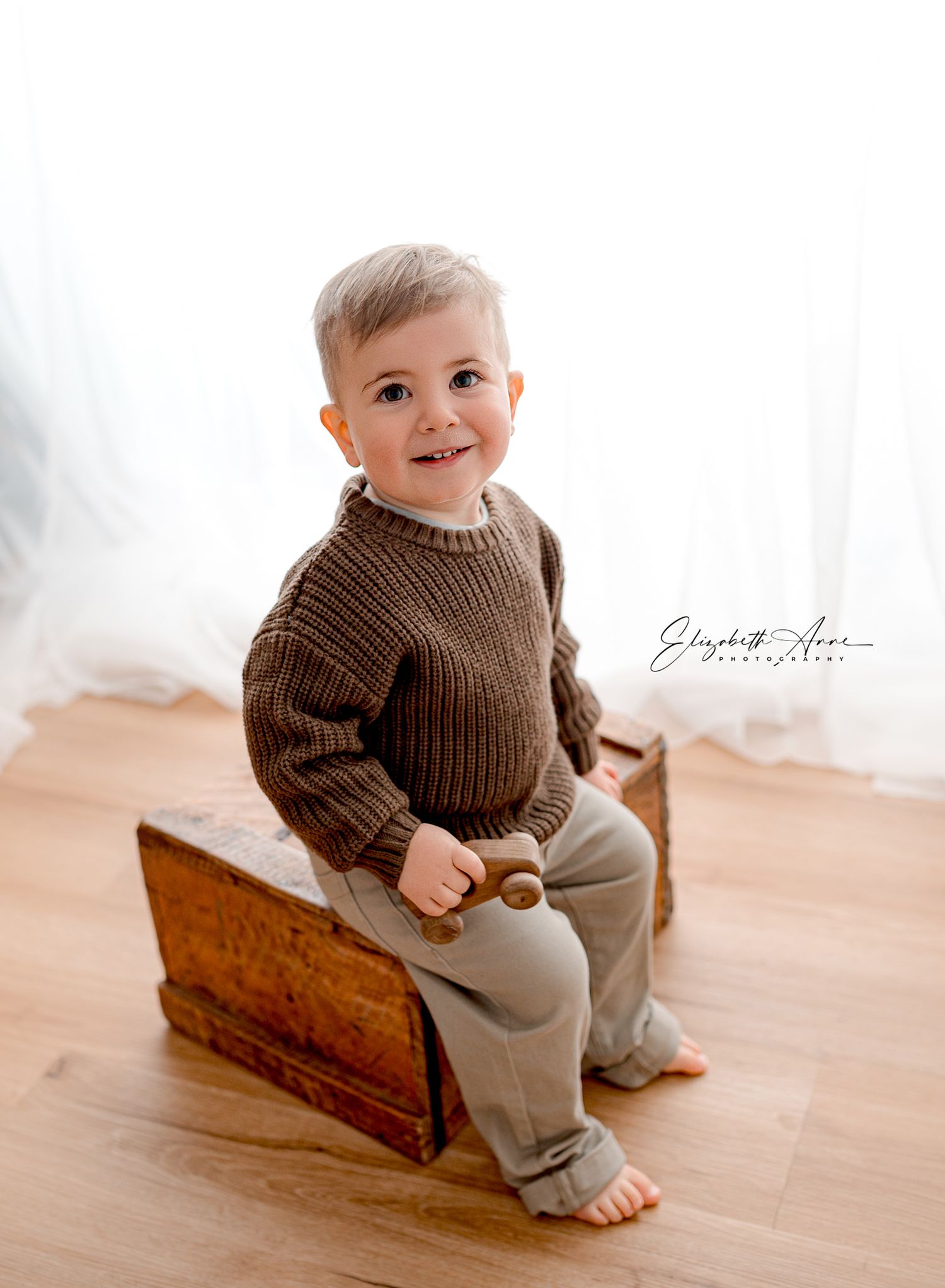 A toddler in a brown knit sweater sits on a vintage wooden box on hardwood floors near white curtains.