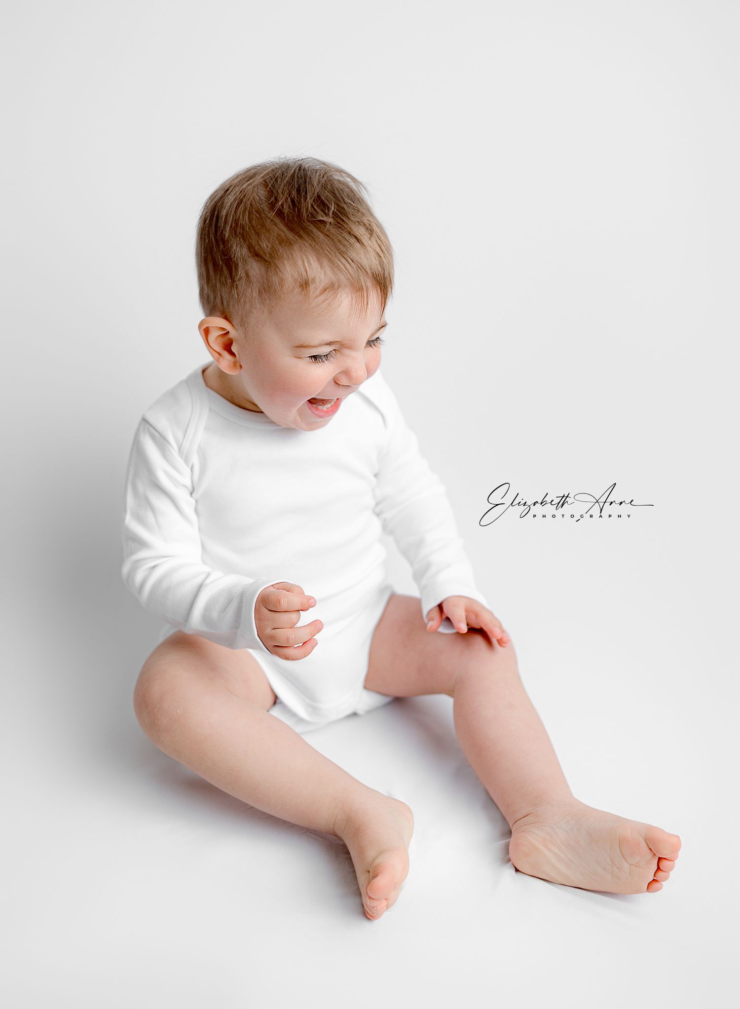 Baby wearing white long sleeve bodysuit laughs playfully while sitting on light gray background during studio photoshoot.