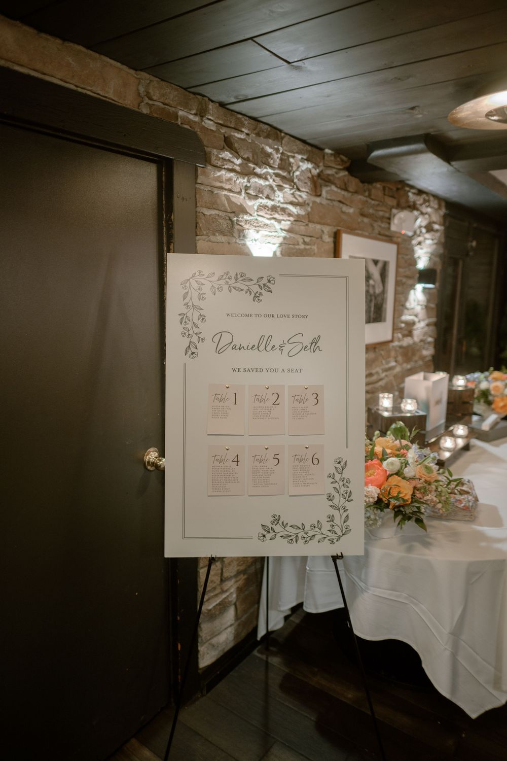 Wedding seating chart displayed on stone wall with floral decorations and white tablecloth in foreground.