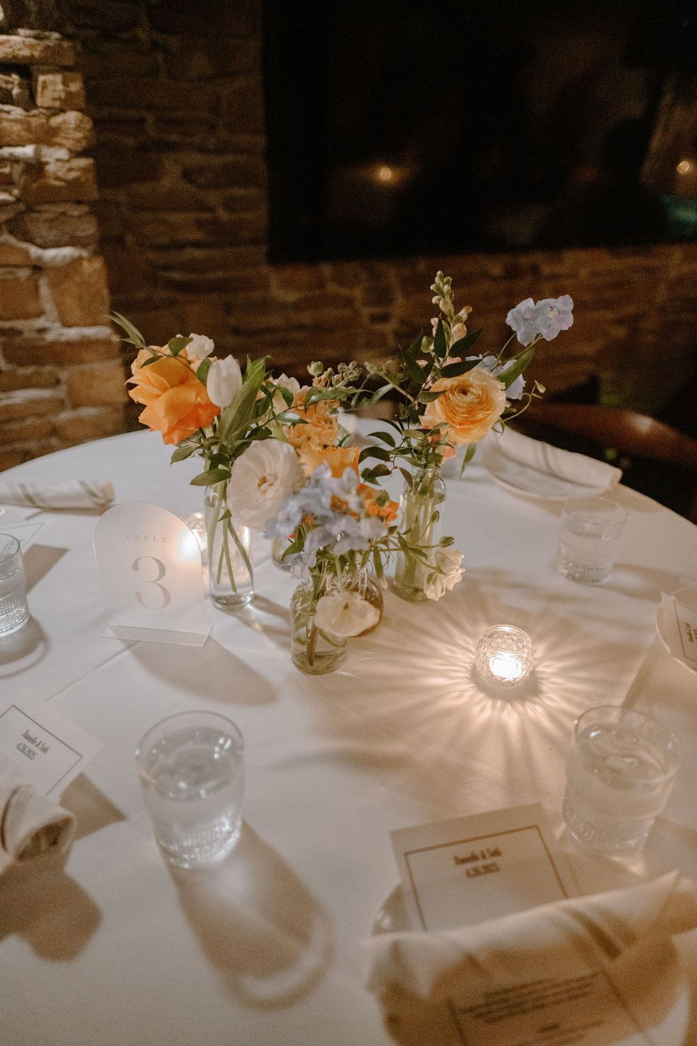 Elegant wedding table setting with peach and white floral centerpiece in mason jar and glowing votive candle.
