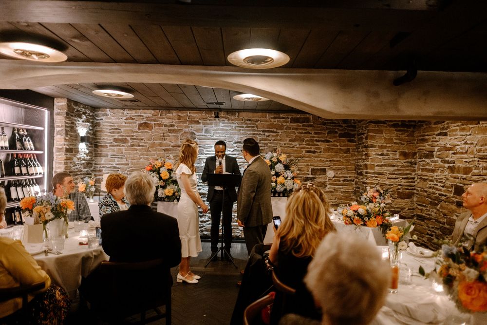 An intimate wedding ceremony takes place in a rustic brick-walled basement venue with warm lighting and autumn decor.