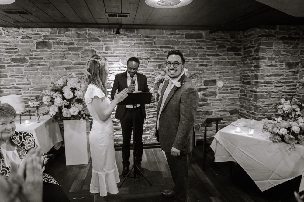 A black and white photograph of an intimate wedding ceremony in a rustic brick-walled venue with floral arrangements.