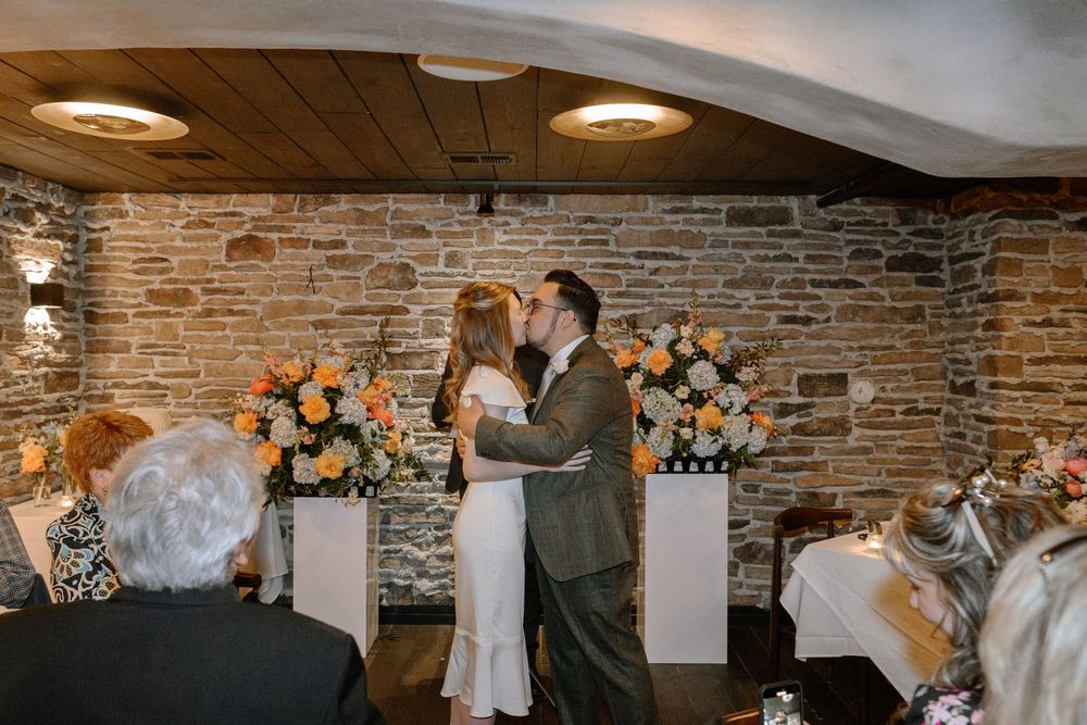A couple shares their first kiss during an intimate wedding ceremony in a rustic brick-walled venue with floral arrangements.