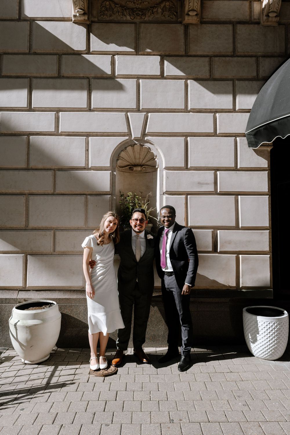 Three people standing in front of white stone building with architectural details and sunlight.
