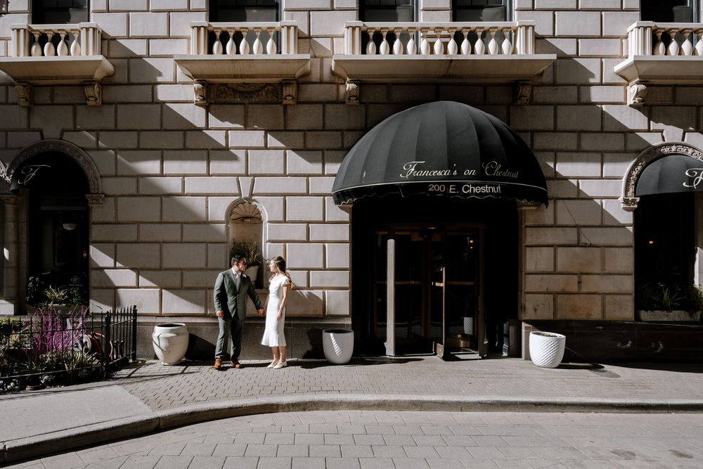Classic stone building entrance with black awnings and white planters along a curved sidewalk.