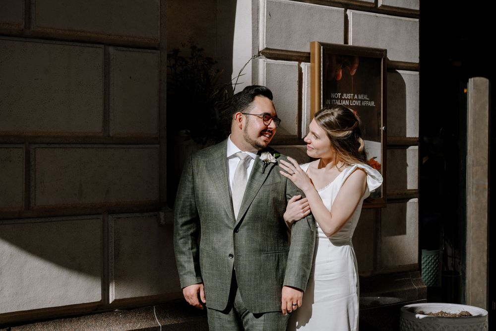 A couple in wedding attire share a romantic moment outside a building in dramatic lighting.