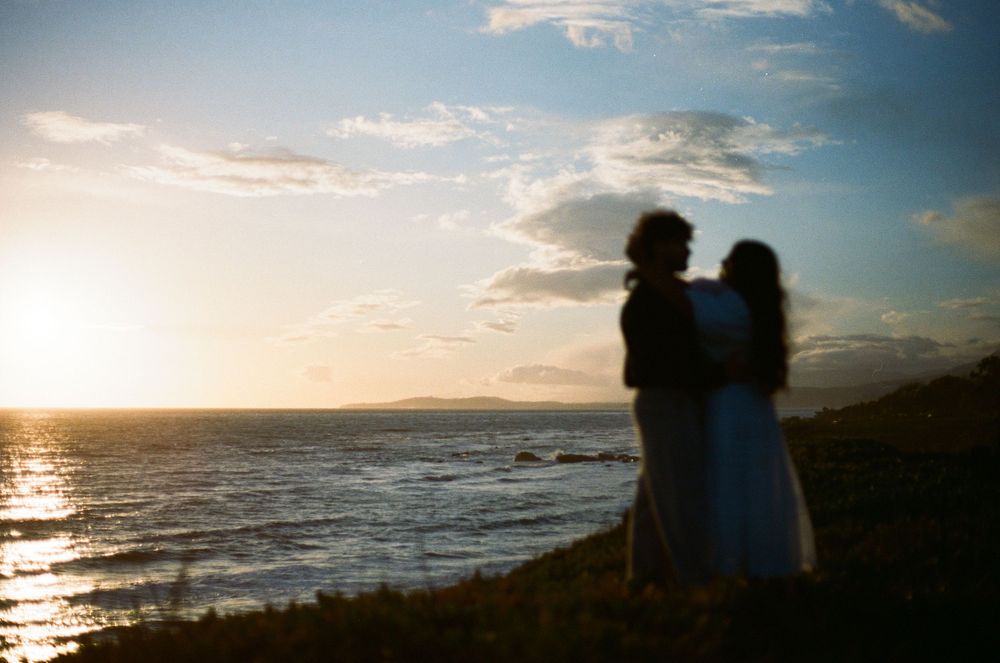 Silhouetted couple standing on coastal cliff overlooking ocean at sunset with dramatic sky and waves.