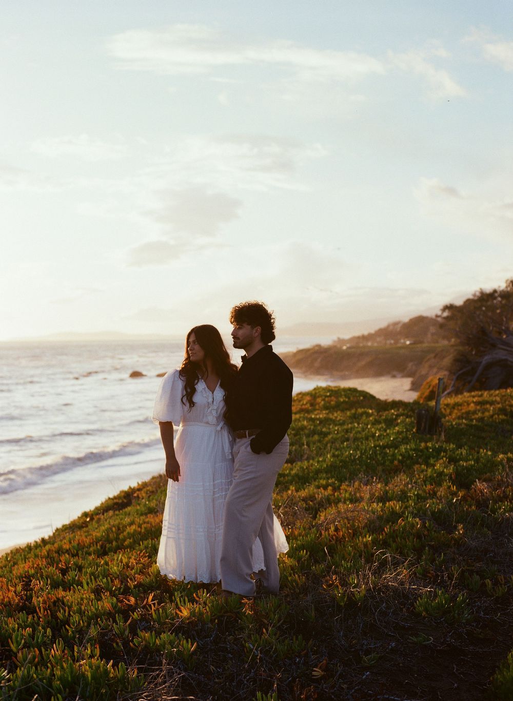 Two people in white clothing stand on a coastal cliff overlooking the ocean at sunset with golden light on grassy terrain.