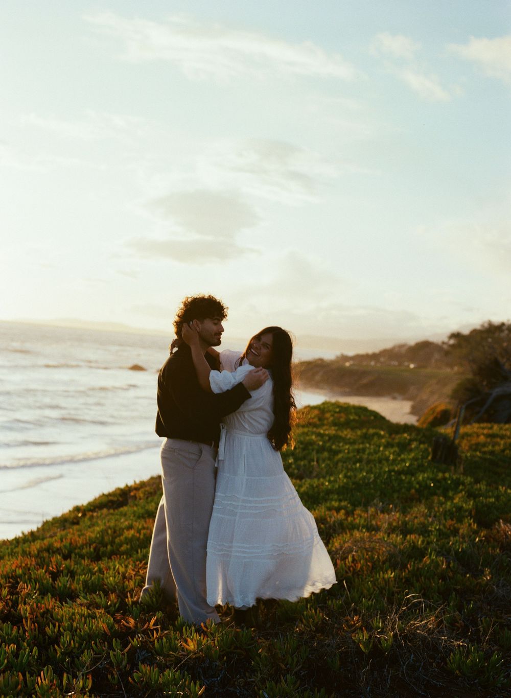 A couple in love embraces on a grassy coastal clifftop at sunset overlooking the ocean and beach below.
