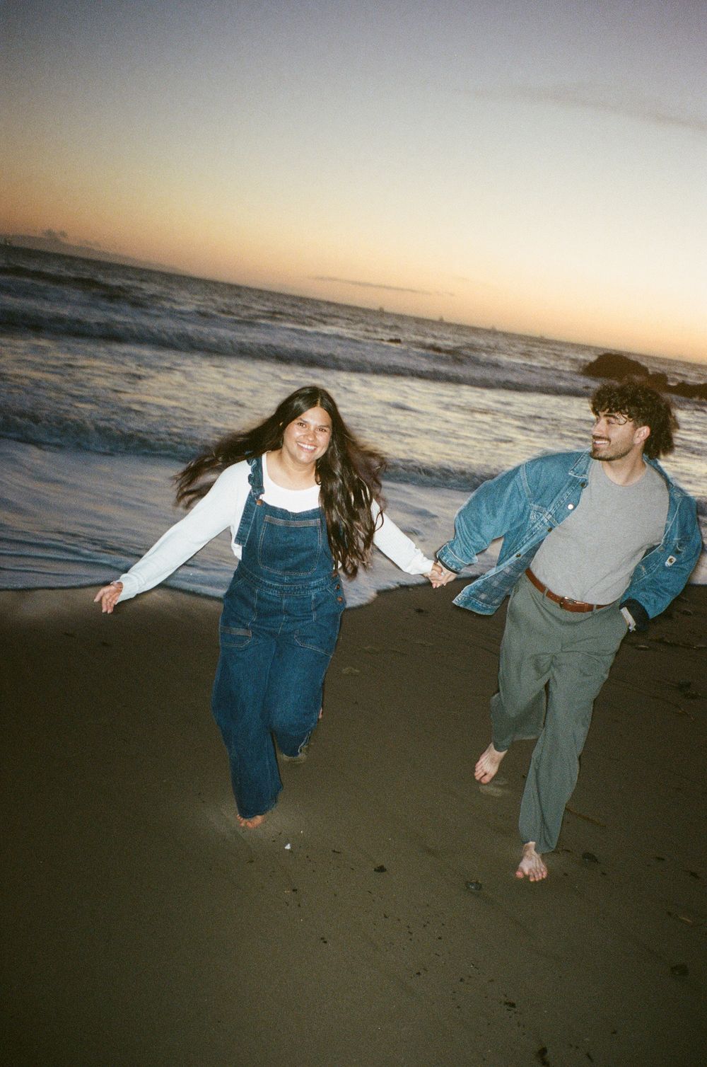 Two people running and holding hands on a beach at sunset with waves crashing in the background.