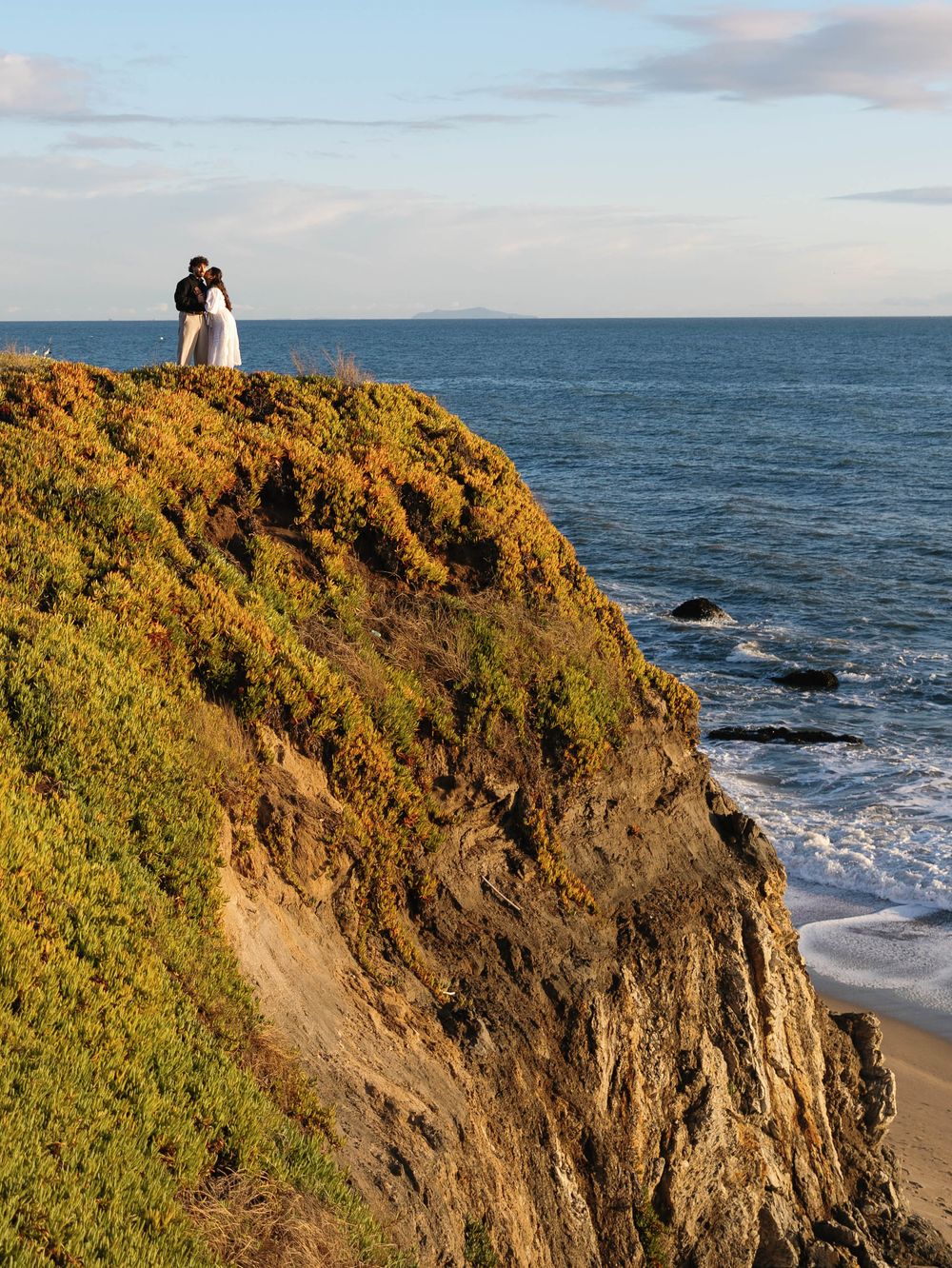 Couple standing on scenic coastal cliff overlooking ocean waves during golden hour at sunset.