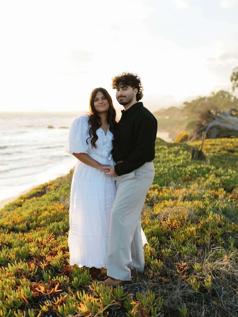 A couple in white outfits embrace while standing on a grassy coastal cliff during golden hour at sunset.