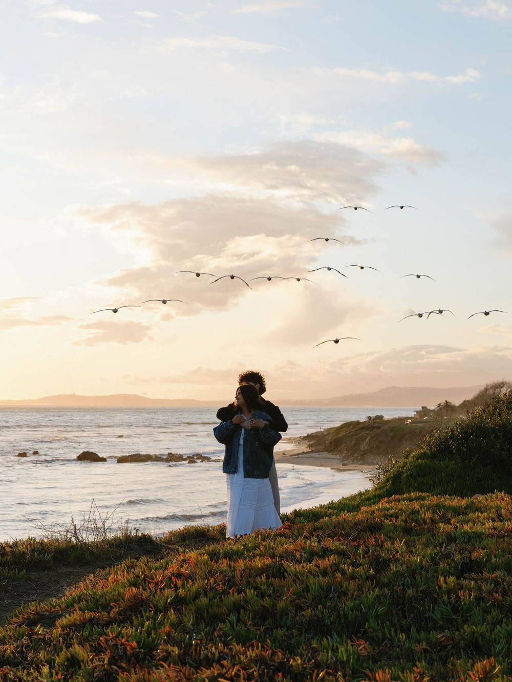 Figure stands on coastal cliff at sunset with birds flying overhead along a dramatic Pacific coastline.