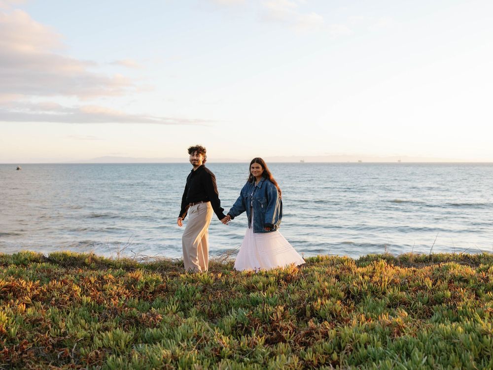 A couple holds hands at sunset on a grassy coastal bluff overlooking the ocean.