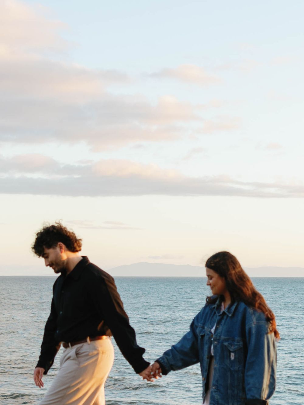 Couple holding hands while walking along the beach at sunset with ocean and cloudy sky in the background.