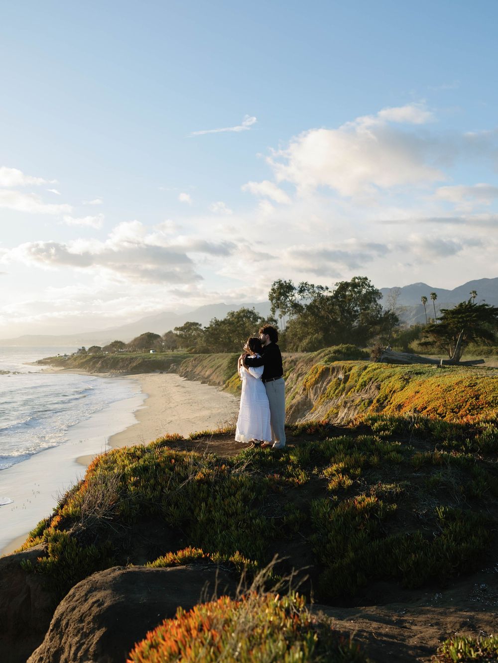 A couple embraces on a scenic coastal cliff at sunset with waves crashing on the beach below and mountains in the distance.