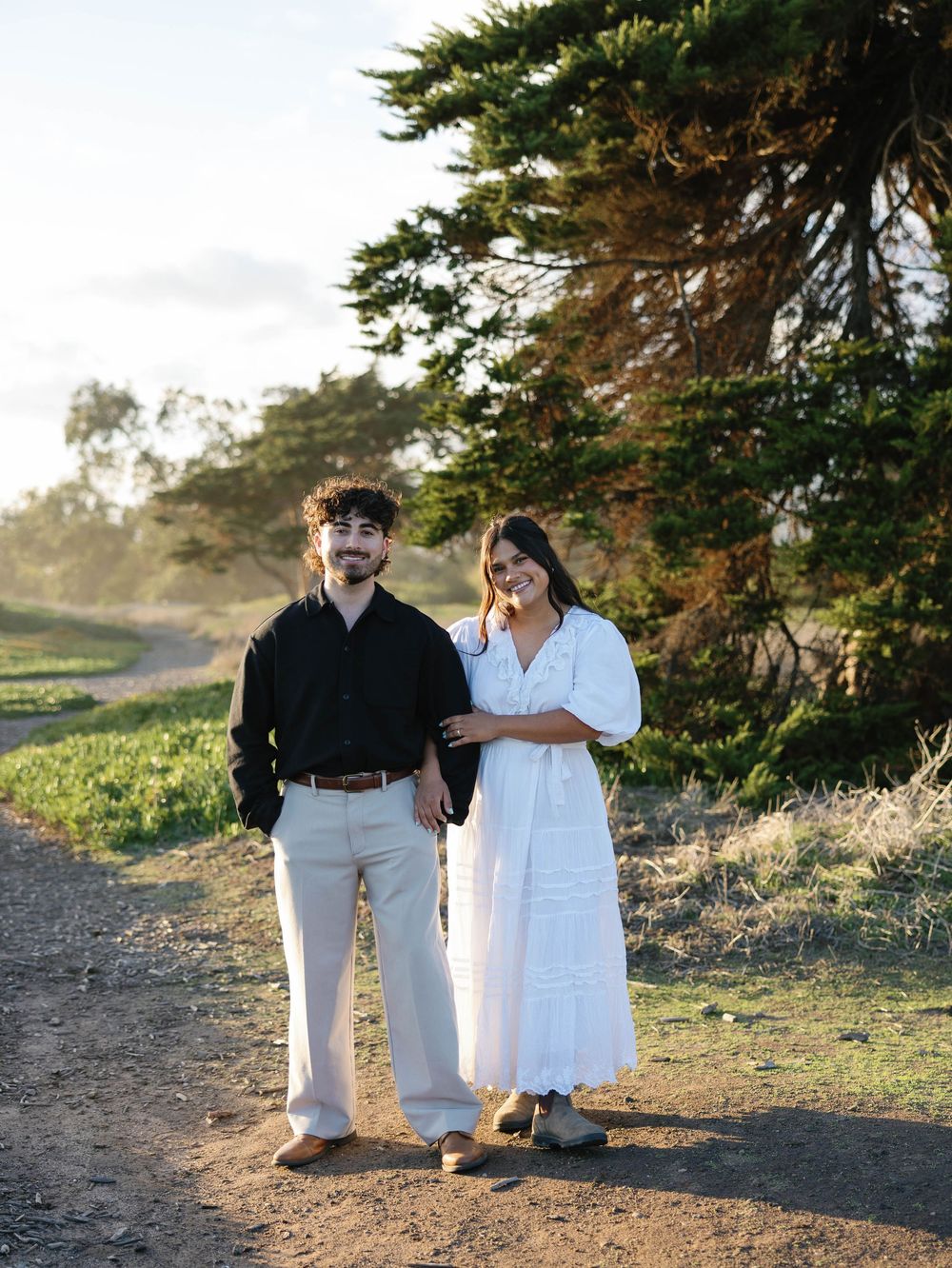 A couple stands together on a dirt path with cypress trees and golden sunlight in the background.