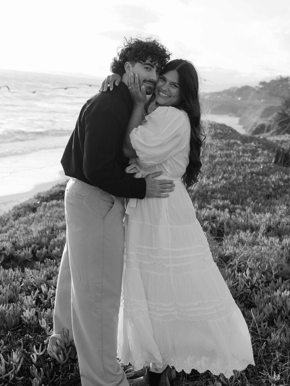 A couple embrace while sharing a joyful moment together on a scenic coastal clifftop overlooking the ocean.