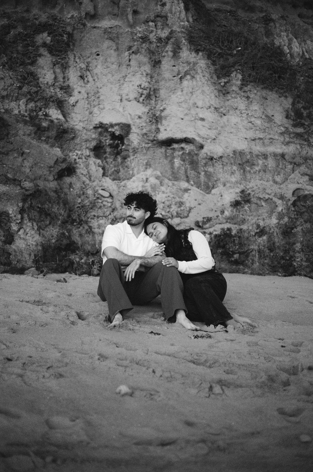 A vintage black and white photograph of two people resting together against a rocky cliff wall at a beach.