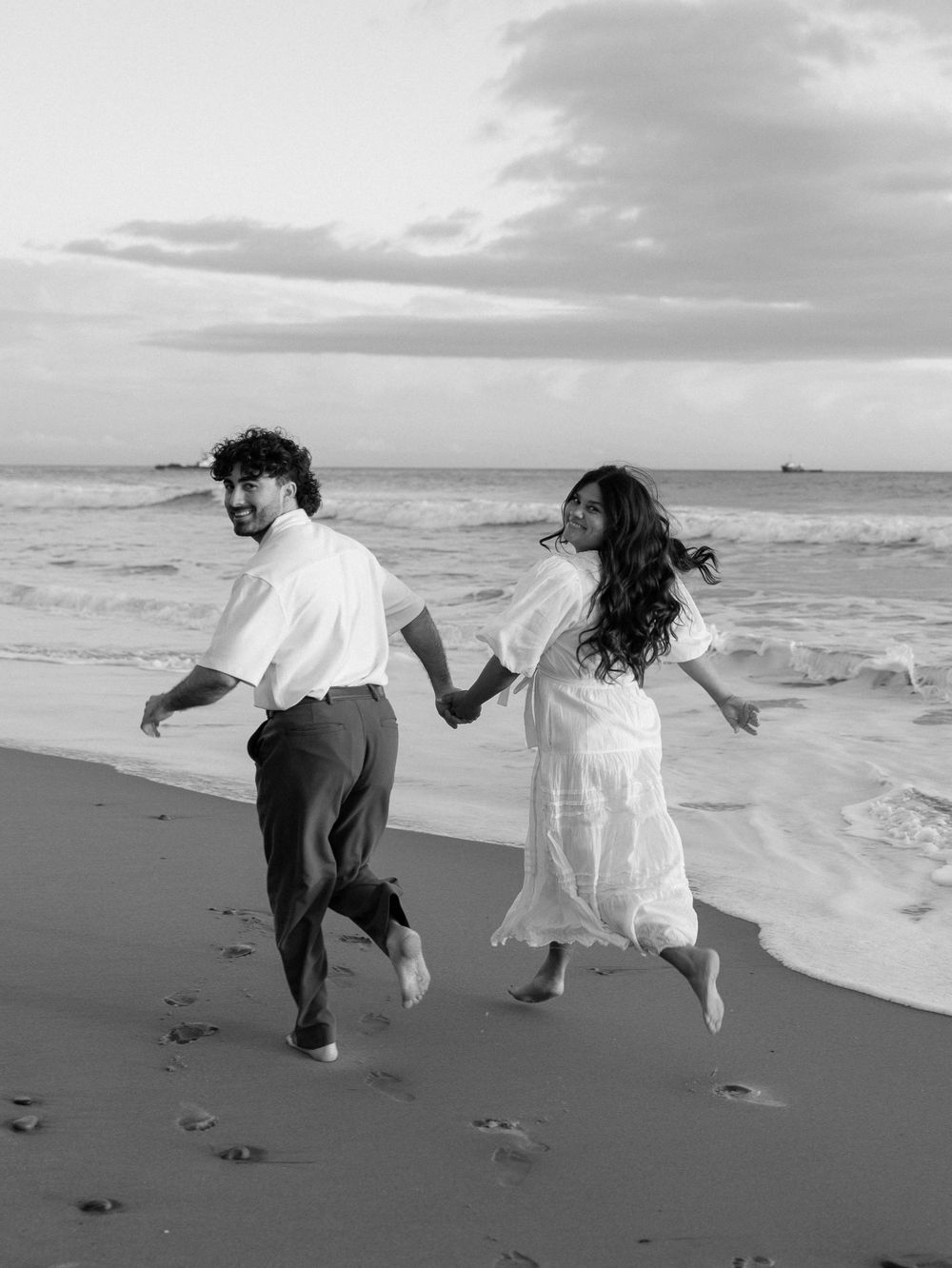A couple runs joyfully together along a beach shoreline in black and white, leaving footprints in the sand at sunset.