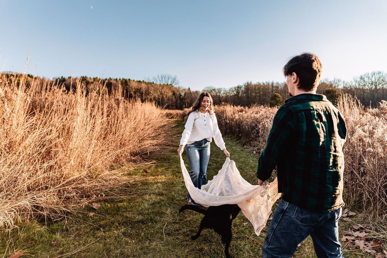 A couple walks through a grassy path surrounded by tall dried cattails and brush on a sunny autumn day.