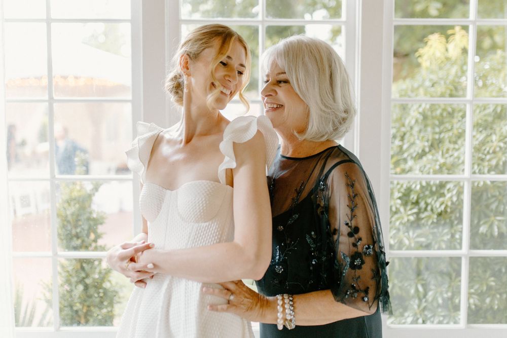 Two people share a joyful moment together by a bright window, with one wearing a white gown and another in black lace.