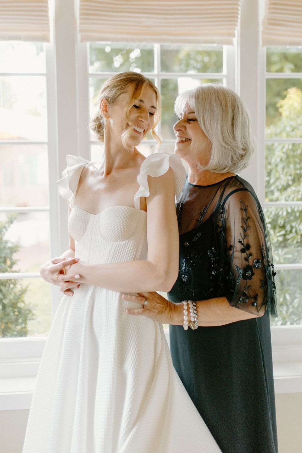 Two women share a joyful moment in formal attire, one wearing a white wedding gown and the other in an elegant black dress.