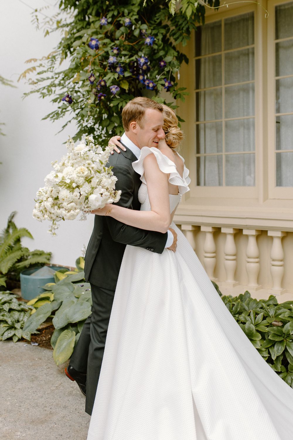 A couple in wedding attire embrace outdoors near a white building with purple flowers and greenery.