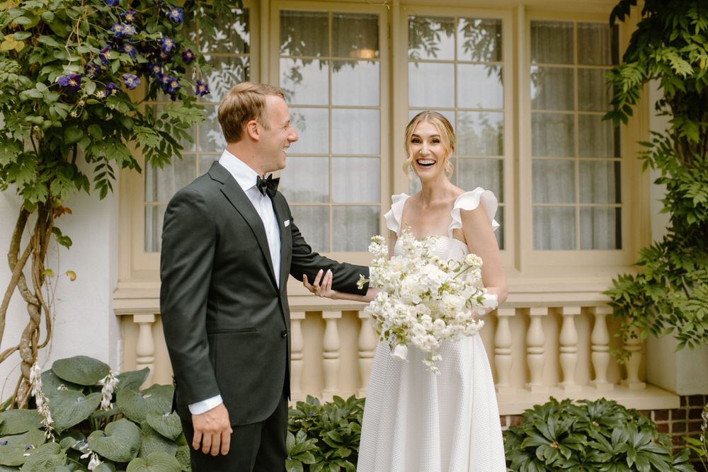 A bride and groom share a joyful moment together on their wedding day outside an elegant home with French doors.