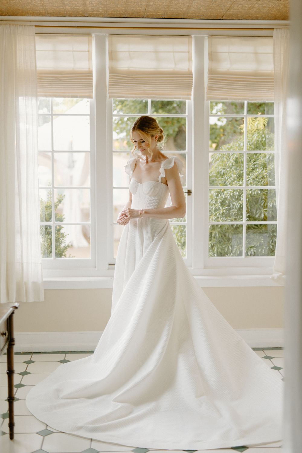 A white wedding dress with a flowing train is elegantly showcased by a window with natural light streaming in.