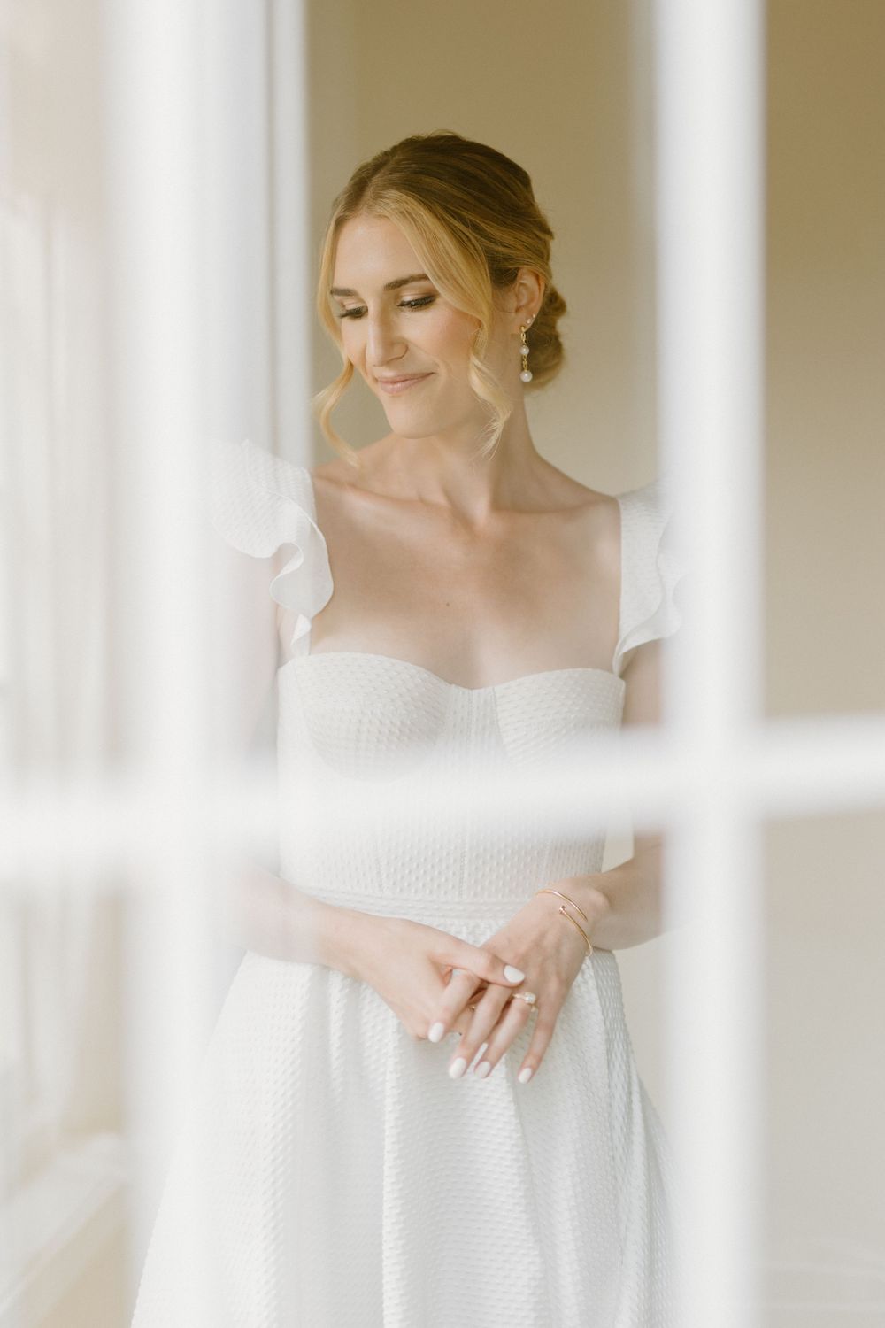 An ethereal bridal portrait through a window frame captures a romantic moment in soft, natural light.