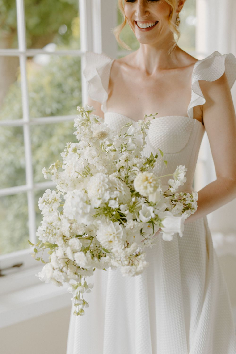 Stunning all-white wedding bouquet with delicate flowers against a light dotted wedding dress with ruffled straps.