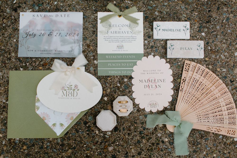 Elegant wedding invitation suite displayed on stone with sage green envelopes, delicate fan, and romantic details.