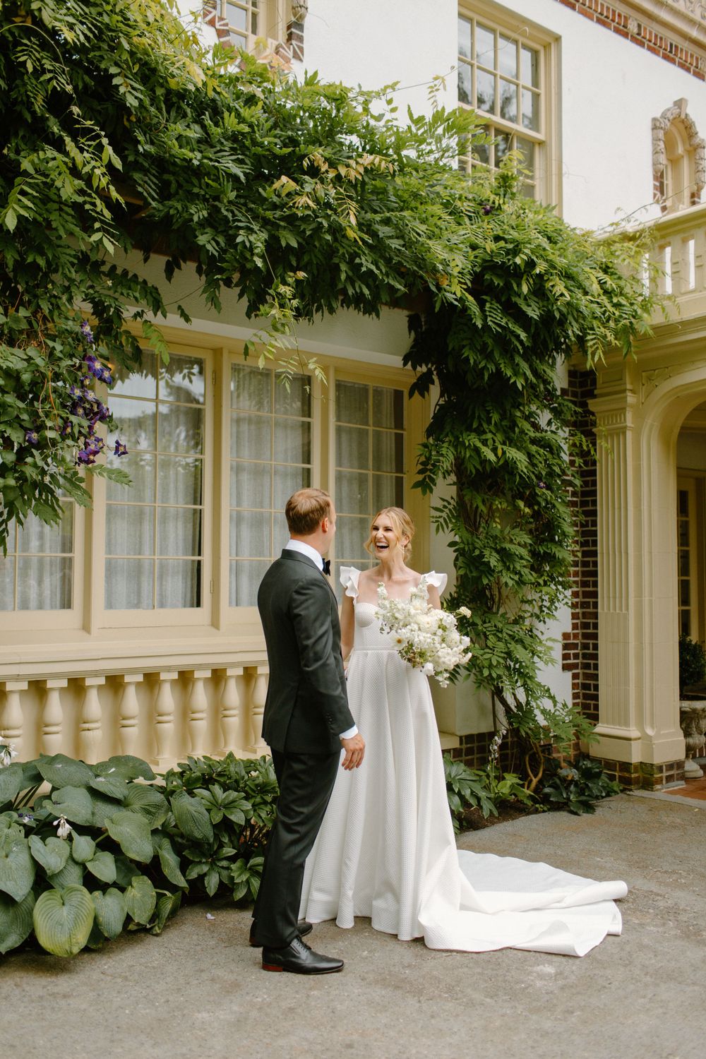 A couple in wedding attire stands beneath lush green vines growing on a classic white stone building exterior.